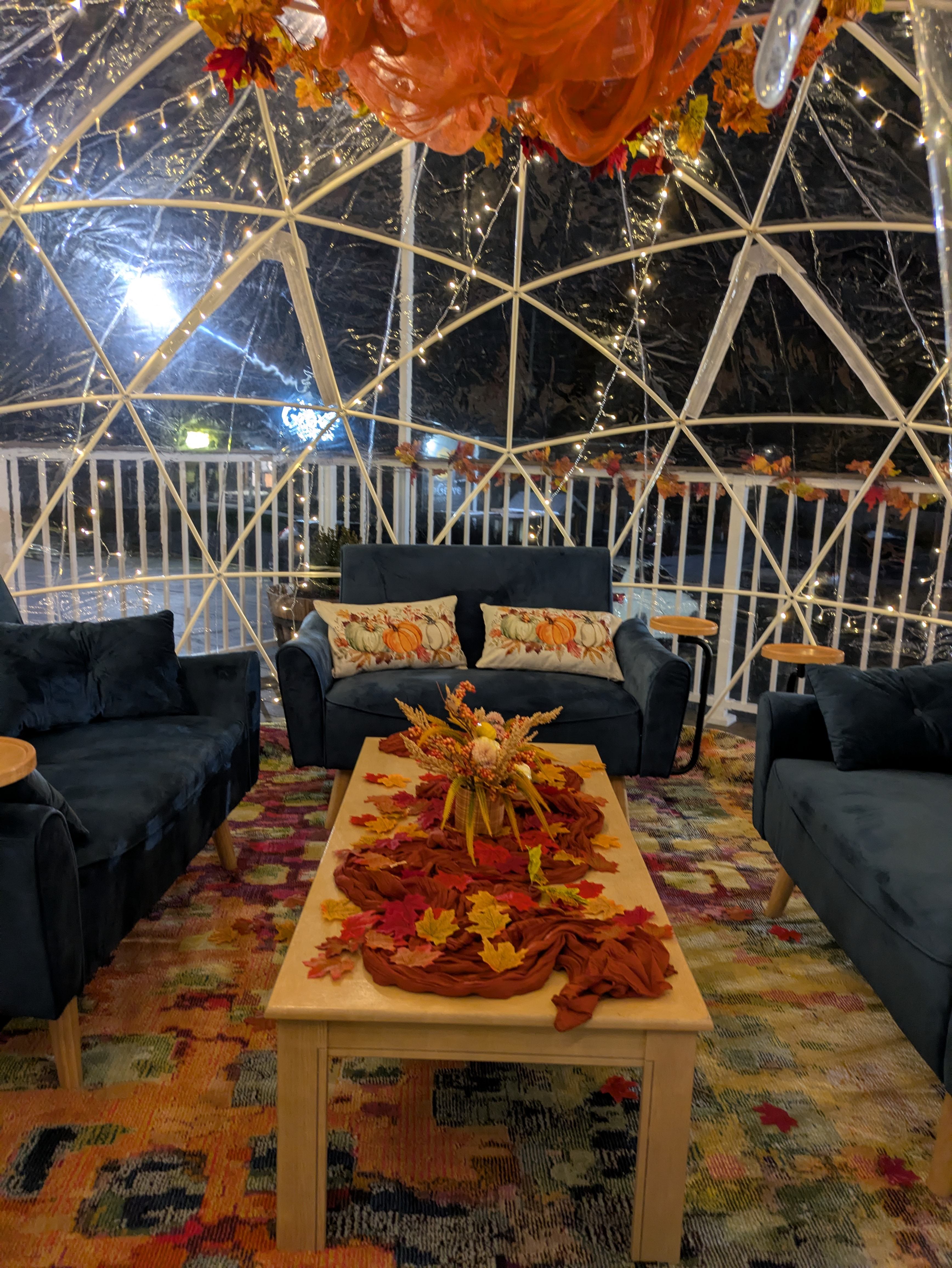 Inside an NYX igloo: blue velvet sofas around a low wooden table dressed with autumn leaves, with string lights across the dome.